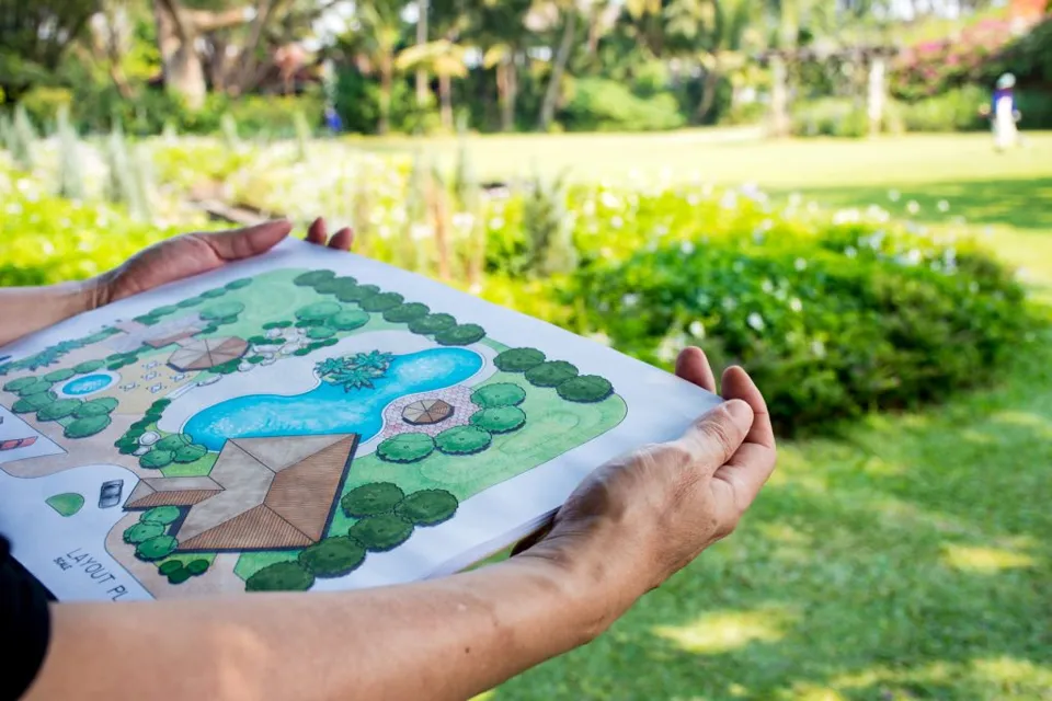 Garden layout plan held in front of a landscaped garden.