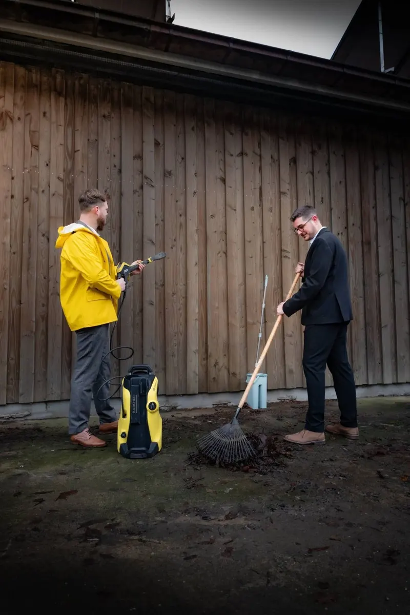 Two people with outdoor cleaning equipment beside a wooden wall.
